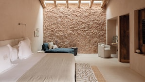 A bedroom with a large bed, a stone wall, a skylight, and a seating area with a chair and a small table. - Hotel Vestige Binidufà (Ferreries)