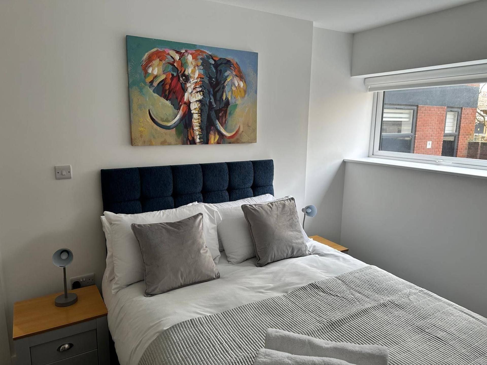 Une chambre à coucher avec un lit, des oreillers, une table de chevet, une lampe et un tableau représentant un éléphant.
