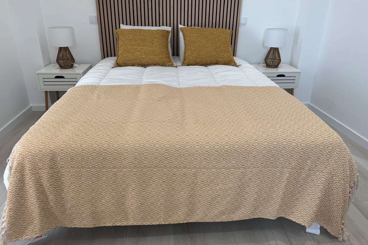 A neatly made bed with a patterned bedspread and two pillows, flanked by white nightstands with lamps and a striped headboard.