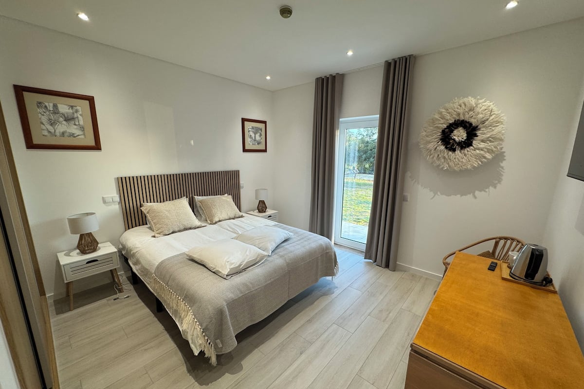 A modern bedroom with a large bed, a bedside table with a lamp, a wooden nightstand, and a view of the outdoors through a sliding glass door.