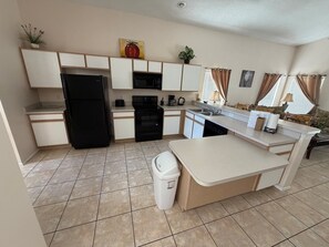 A compact kitchen with a refrigerator, microwave, oven, and dishwasher. There is a small trash bin and a window with curtains.
