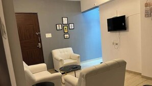 Ein modernes Zimmer mit einer Couch, Sesseln und einem kleinen Tisch. Eine Tür, ein an der Wand befestigter Fernseher und gerahmte Bilder sind vorhanden.