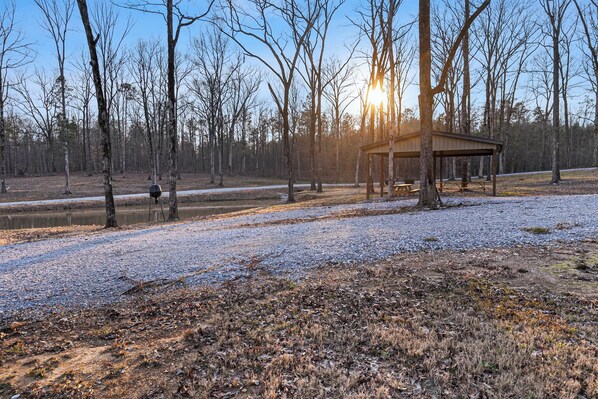 A covered pavilion in a forested area with a gravel path leading to a body of water.