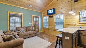 Un salón acogedor con techo de madera, un televisor montado en la pared y una zona de cocina con encimera y armarios.
