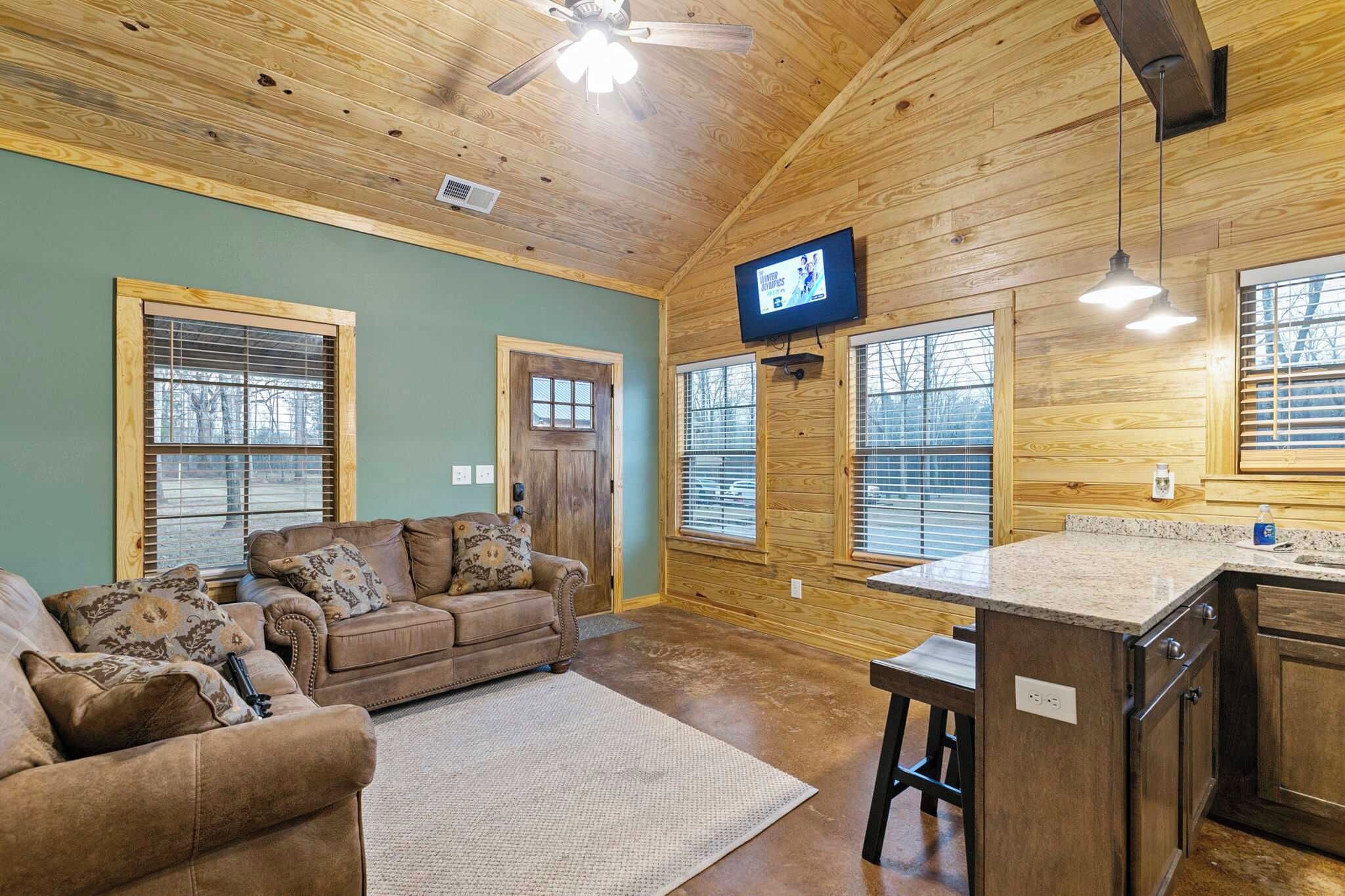 A cozy living room with a wooden ceiling, a television mounted on the wall, and a kitchen area with a countertop and cabinets.