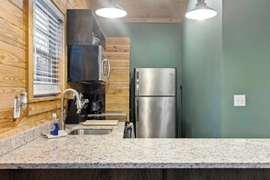 Una cocina con encimera de granito, refrigerador de acero inoxidable, armarios de madera y una ventana con persianas.