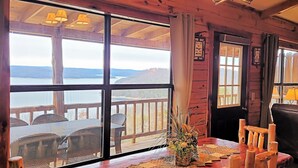 Interior acogedor de una cabaña con grandes ventanales que dan a un lago y montañas, una chimenea y muebles de madera rústica.