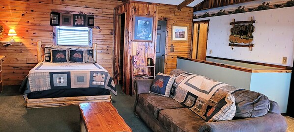 A cozy cabin-style room with a wooden bed, a couch, and a wooden bench. There are framed pictures on the walls and a lamp on a side table.