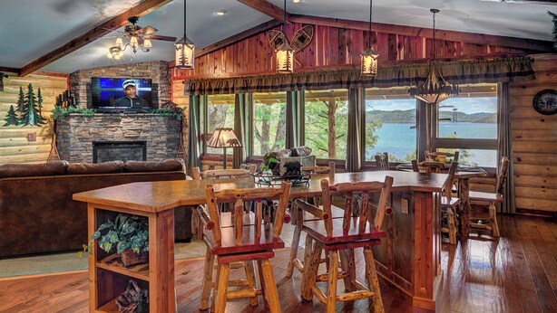 A rustic cabin interior with a stone fireplace, wooden furniture, and a large window overlooking a lake.