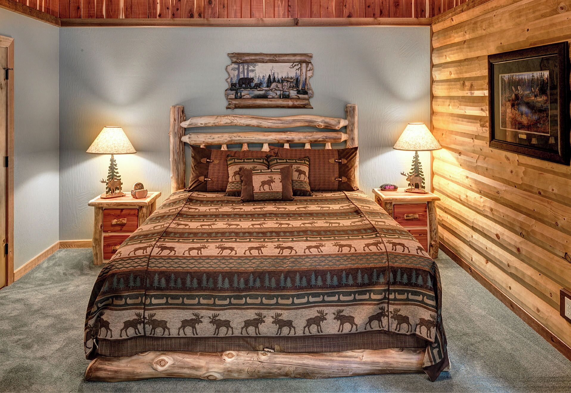 A log cabin-style bedroom with a bed centered between two nightstands, each with a lamp. The walls are adorned with wooden paneling and a framed picture above the bed.