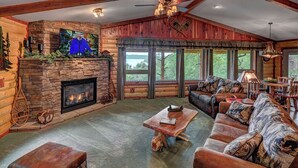 A cozy log cabin interior with a stone fireplace, leather furniture, and large windows offering a view of the outdoors.
