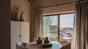 A room with a round table, two chairs, a book, and two vases. There is a window with curtains, and a view of a beach with lounge chairs. - Hotel Vestige Son Ermità (Ferreries)
