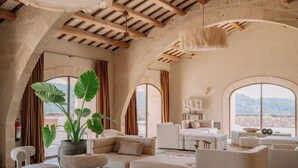 A spacious living room with a high ceiling, stone walls, and large arched windows offering a view of the mountains. - Hotel Vestige Son Ermità (Ferreries)