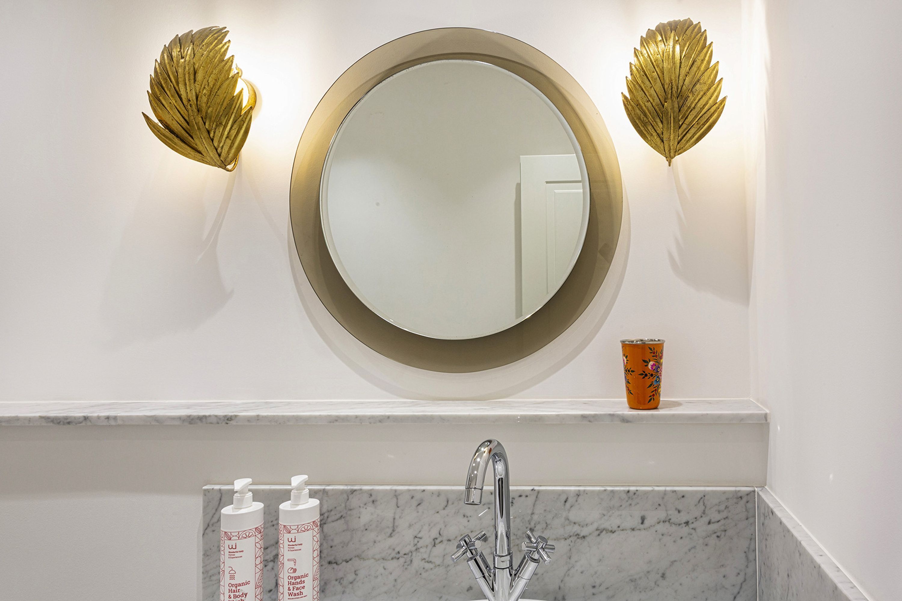 Une salle de bain avec un miroir rond, deux appliques murales, un lavabo et deux bouteilles de gel douche biologique.