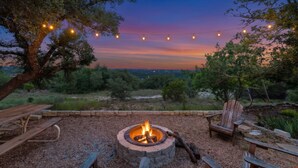 A campfire surrounded by wooden benches and a picnic table, with string lights overhead and a view of the landscape. - Beautiful French Country Villa wt Pool & Fire-pit in marvelous Austin (Austin)
