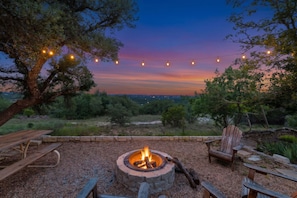 Una fogata rodeada de bancos de madera y una mesa de picnic, con luces colgantes y vistas al paisaje.