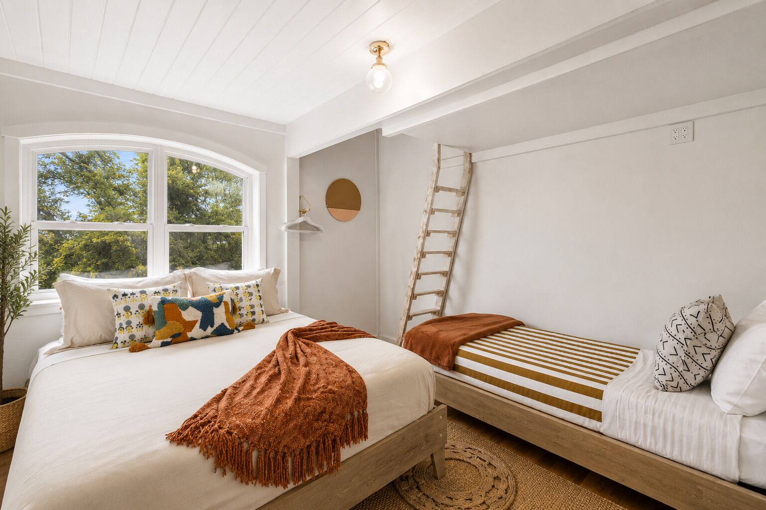 A bedroom with a large window, a bed with patterned pillows, a smaller bed with striped bedding, a ladder, and a hanging light fixture.
