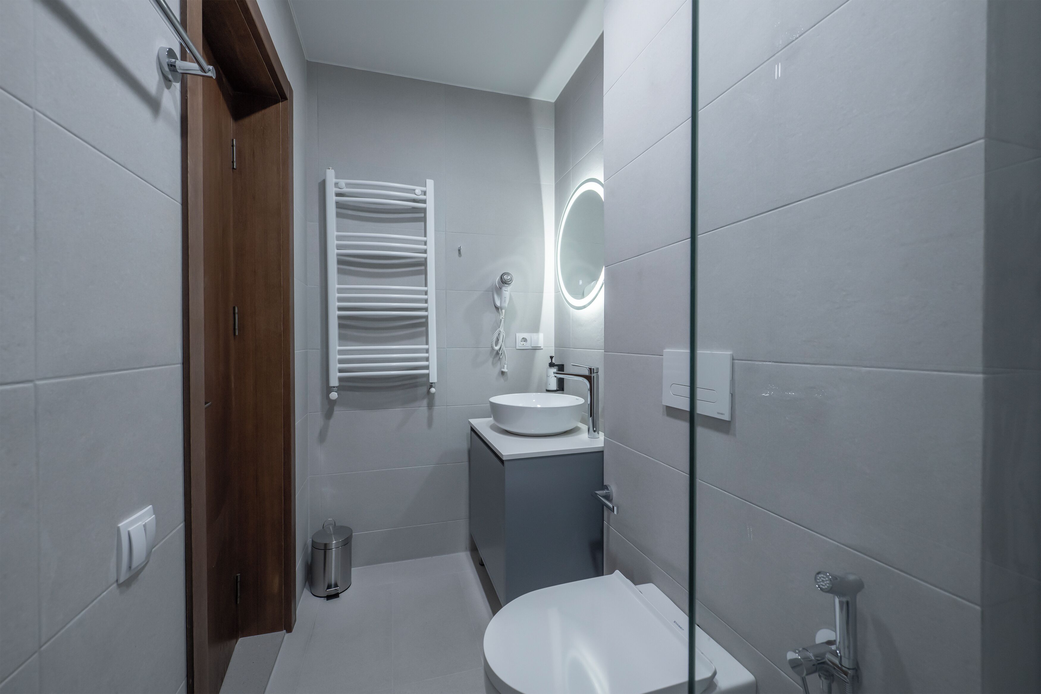 A modern bathroom with a white sink, a mirror with a circular light, and a towel rack.