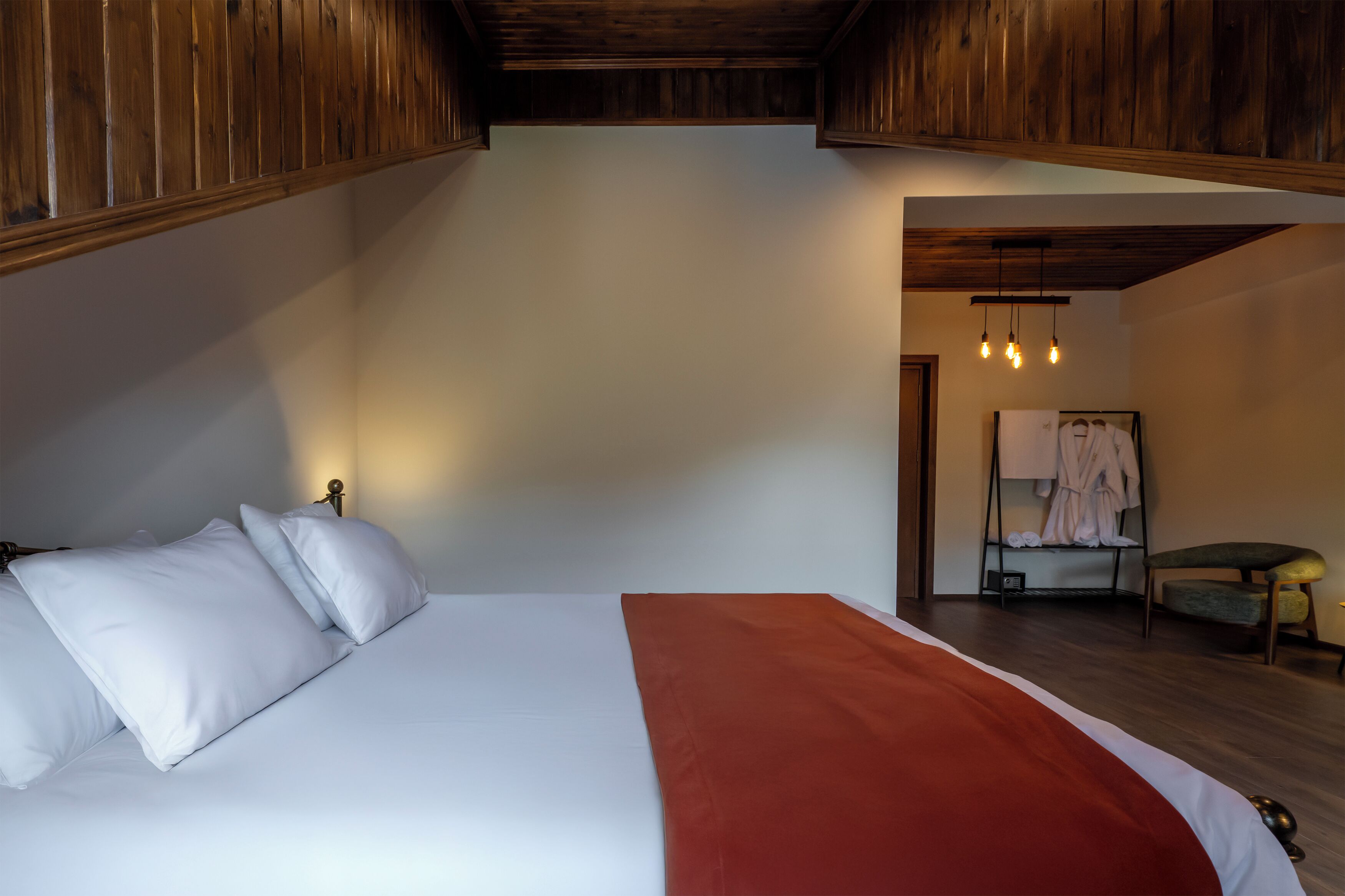 A bedroom with a bed, white pillows, a red throw blanket, wooden ceiling beams, a small chair, and a clothing rack with a towel.