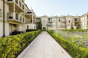 A pathway flanked by neatly trimmed hedges and lavender bushes, leading to a row of beige multi-story residential buildings with balconies. - Corte Fasanella 50 (Toscolano-Maderno)