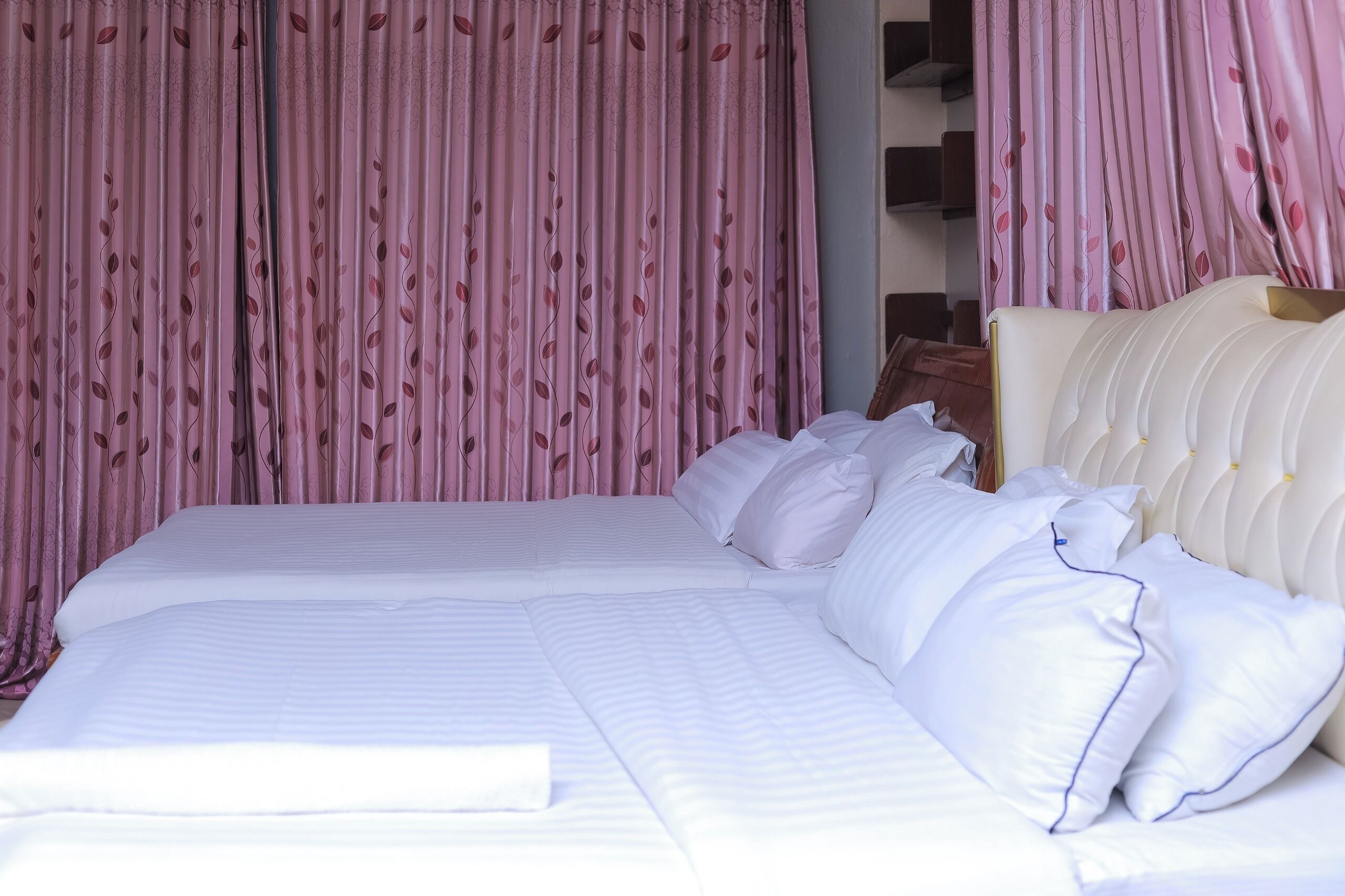 A neatly made bed with white linens and pillows, a white headboard, and pink curtains with a leaf pattern.