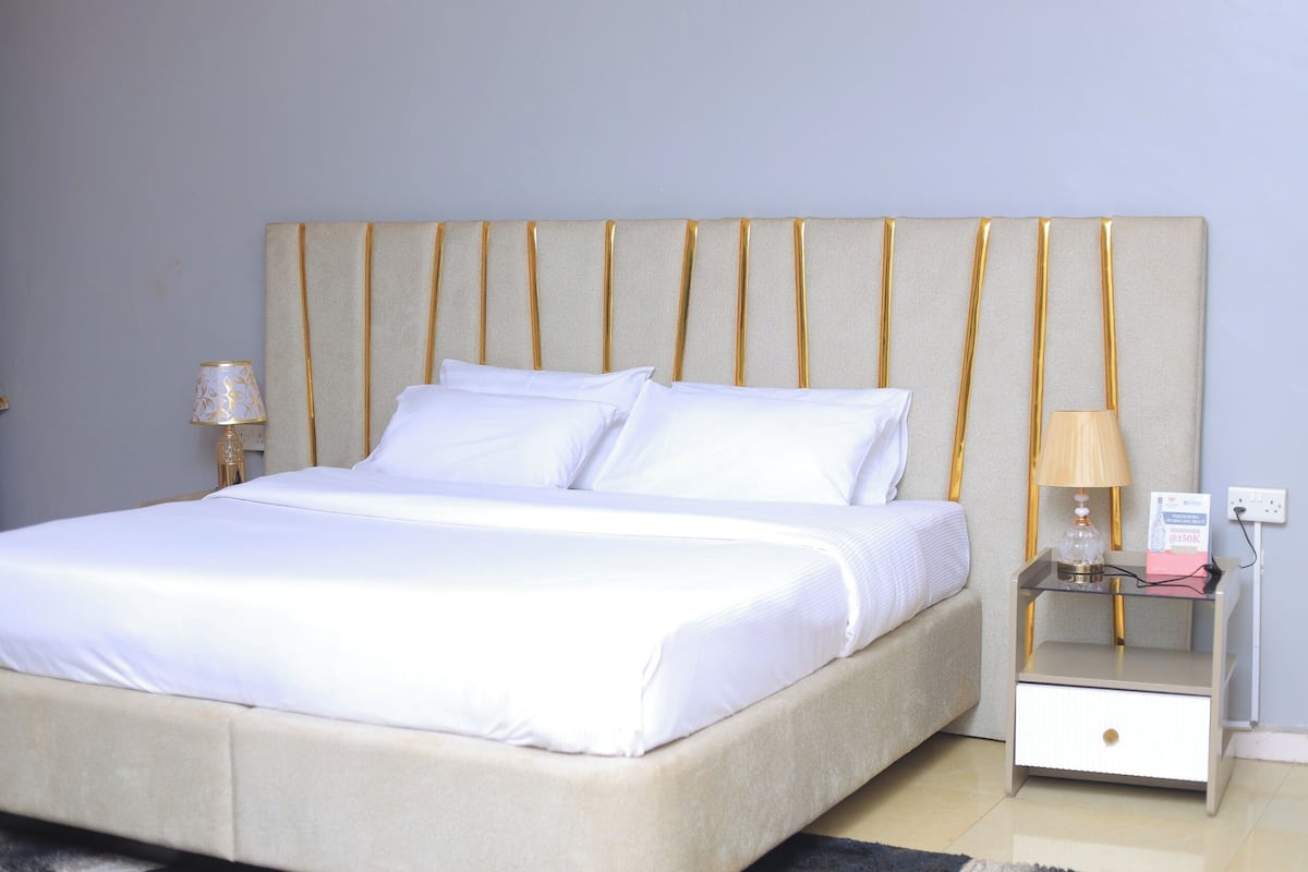 A neatly made bed with white linens, a headboard with vertical gold accents, and a bedside table with a lamp and a power outlet.