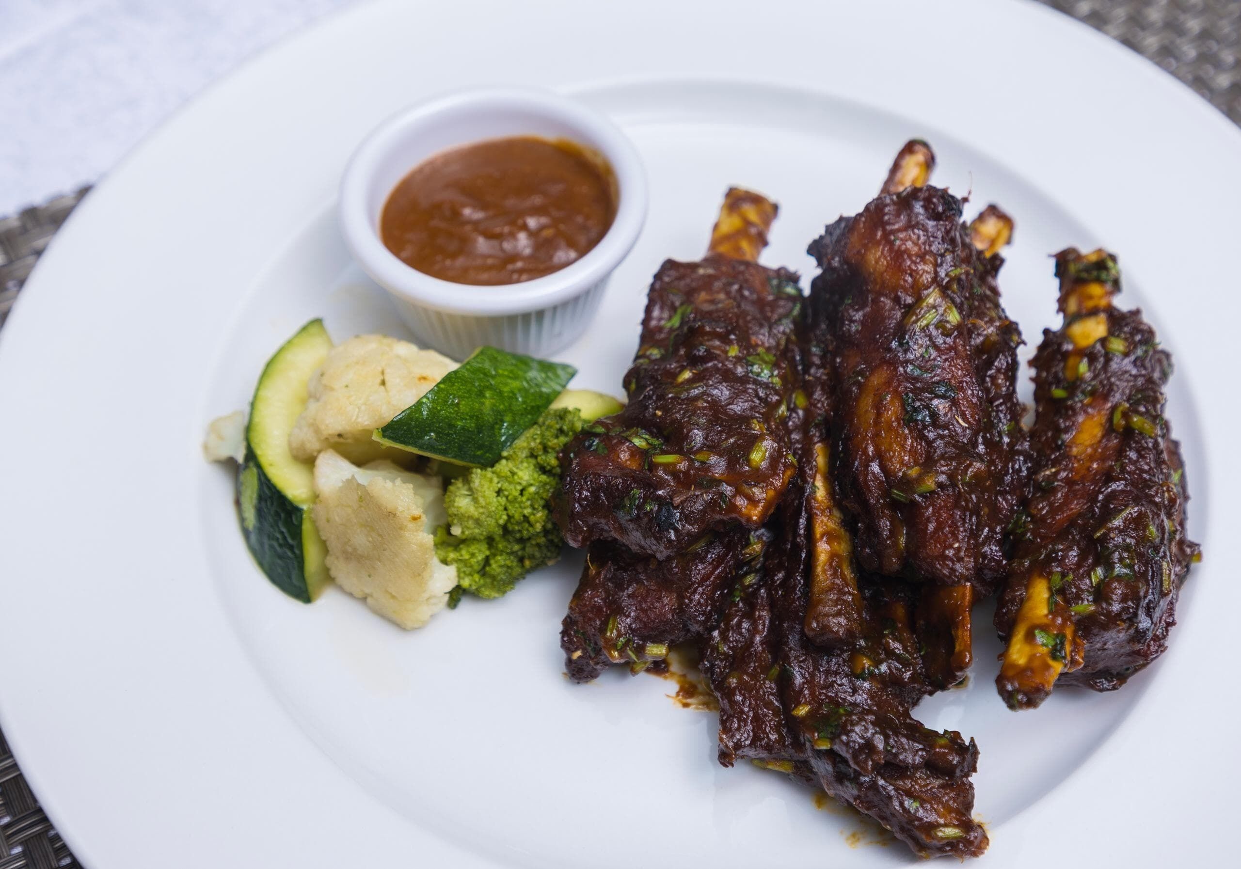 A plate of grilled ribs with a side of sauce and vegetables.
