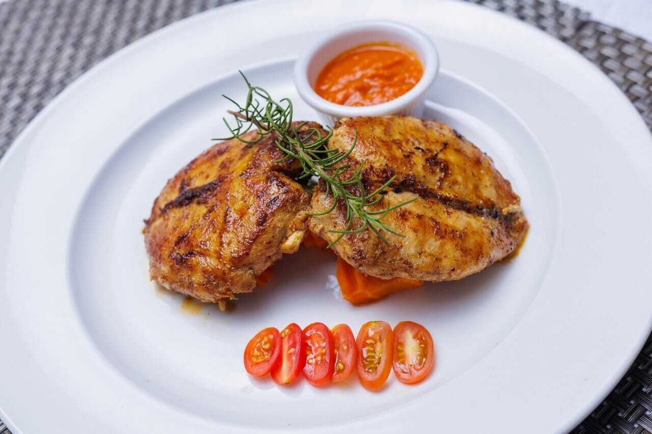 A plate with grilled chicken, a small bowl of sauce, and sliced tomatoes.