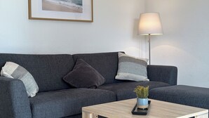 Un salon moderne avec un canapé gris, un tableau encadré représentant une plage, une lampe sur pied et une petite table sur laquelle est posée une plante.