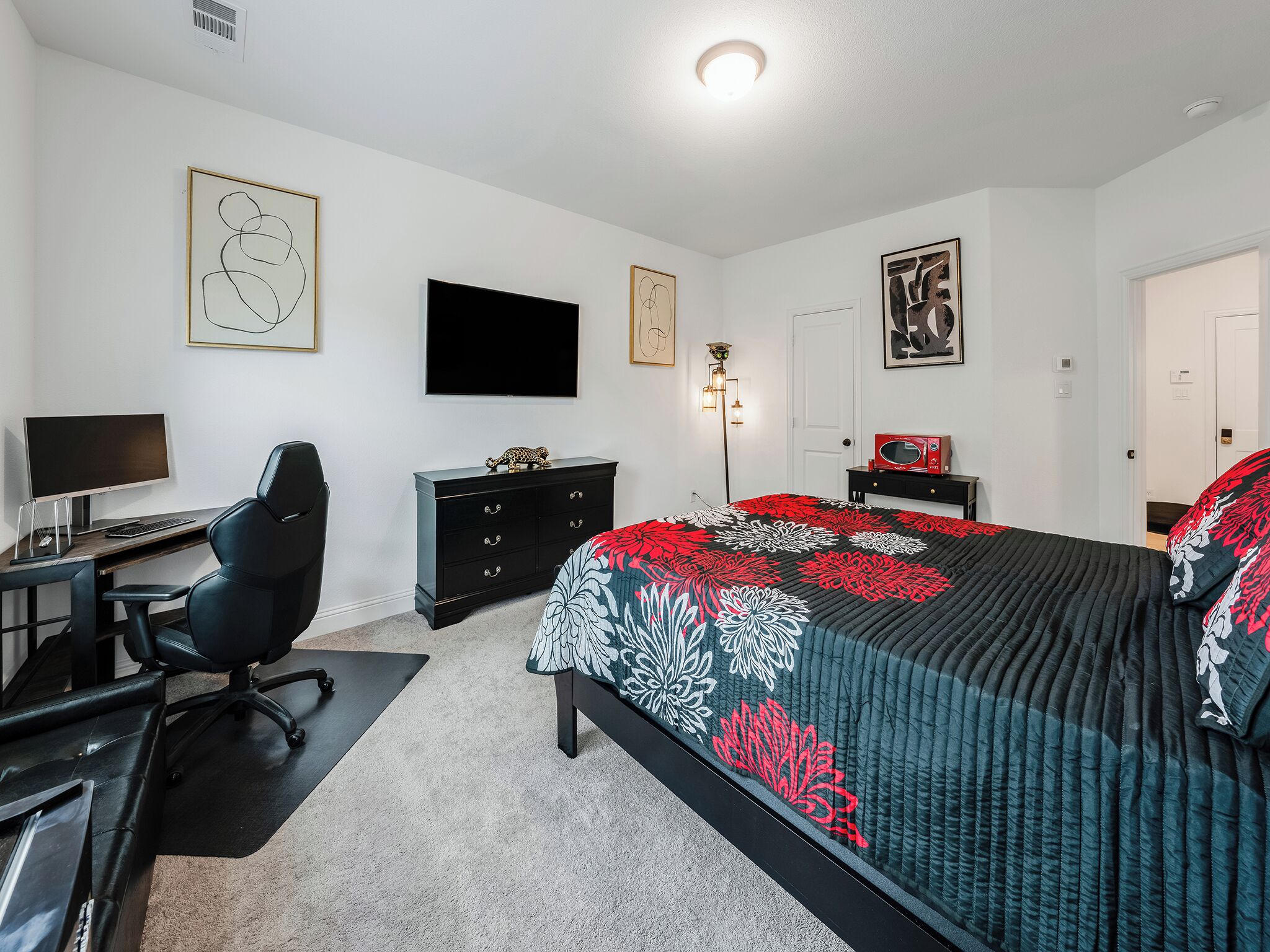 A bedroom with a bed, a desk with a computer, a chair, a dresser, and a TV on the wall.