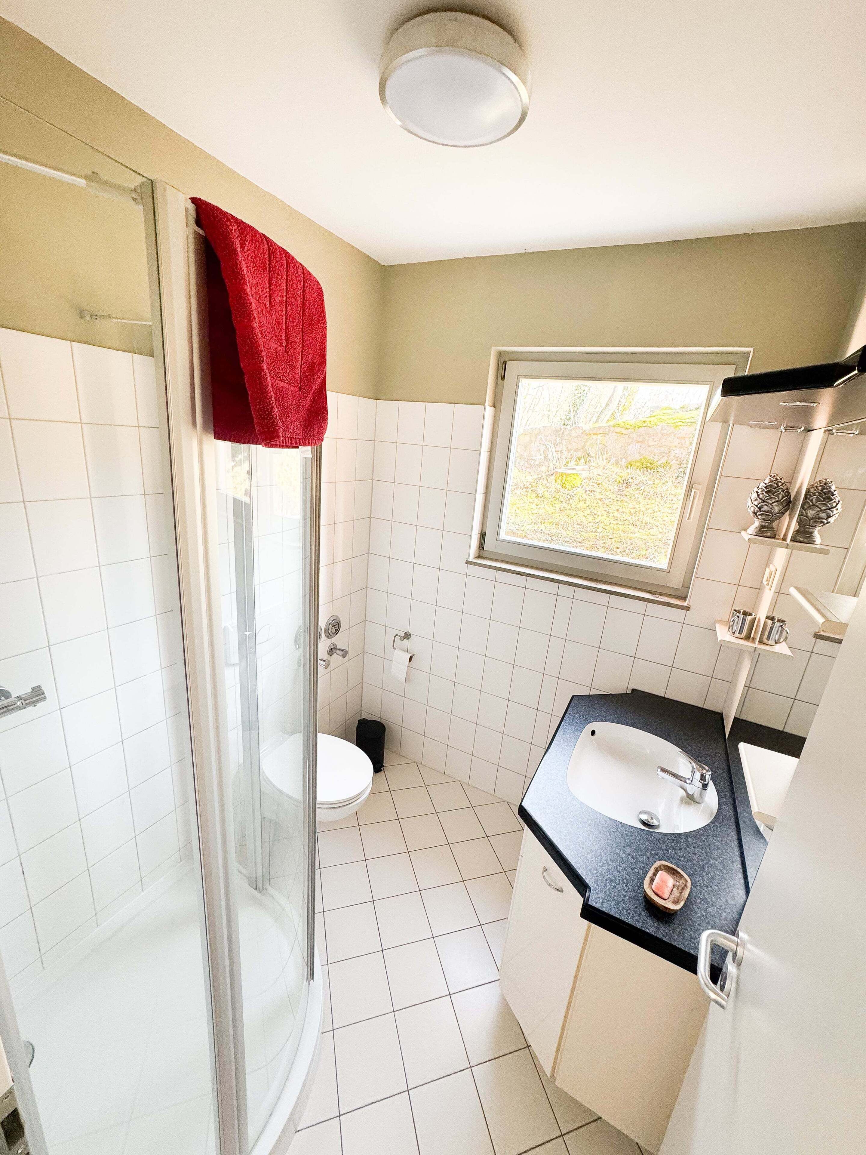 Ein Badezimmer mit einer Duschkabine, einer Toilette, einem Waschbecken mit schwarzer Arbeitsplatte und einem Fenster mit Blick ins Grüne.