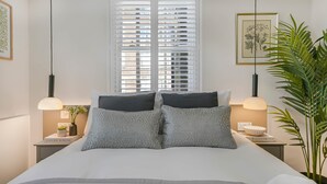 A bedroom with a bed, two bedside tables, a window with blinds, a hanging light fixture, and a potted plant. - Studio 9 @ OneStay Maida Vale (London)