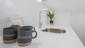 A kitchen with a white countertop, a stainless steel sink, a potted plant, and two mugs with the text 'onestay'. - Studio 18 @ OneStay Maida Vale, Studio 18 at One Stay (London)