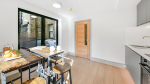 A modern kitchen with a wooden table, bar stools, a large window, and a door leading to another room. - Studio 20 @ OneStay Maida Vale (London)