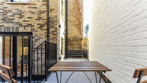 A narrow alleyway with a wooden table and benches, brick walls, and a white wall with a window. - Studio 20 @ OneStay Maida Vale (London)