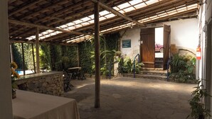 An outdoor patio with stone flooring, wooden tables, and chairs. There is a stone wall with a wooden door, and the area is surrounded by lush greenery. - Suite Confort 201 (Centro Diamantina)