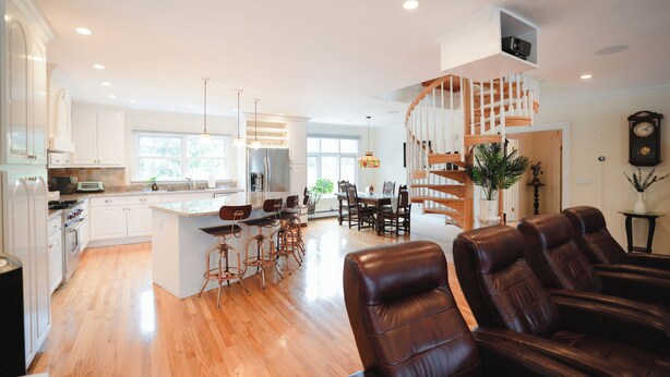 A spacious kitchen with wooden flooring, a central island, and modern appliances. There is a dining area with a chandelier and a staircase leading to an upper level. - Spacious Waterview Home - Spa, Pool Table & Fireplaces – for Families & Reunion (Bourne)