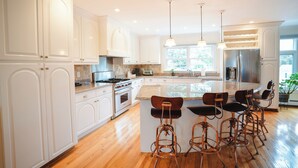 A modern kitchen with white cabinetry, a central island with bar stools, stainless steel appliances, and pendant lighting. - Spacious Waterview Home - Spa, Pool Table & Fireplaces – for Families & Reunion (Bourne)
