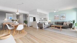 A modern living room with a grey sofa, a wooden coffee table, and a dining area with a wooden table and white chairs.