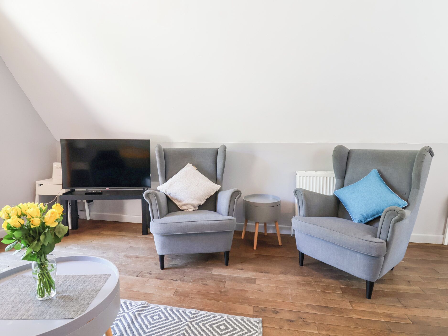 Una sala moderna con dos sillones grises, una mesita auxiliar, un televisor de pantalla plana y un jarrón con flores amarillas sobre una mesa redonda.