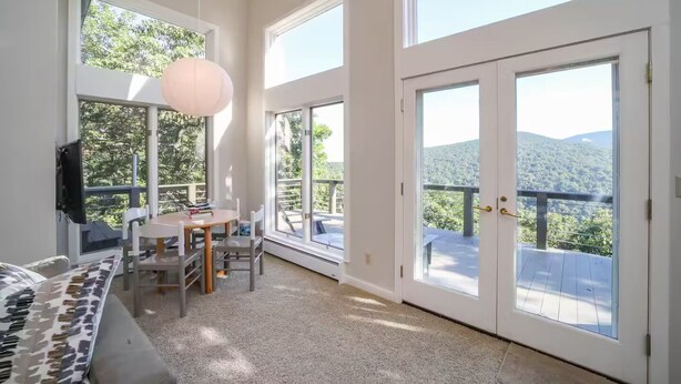 A spacious room with large windows offering a view of a forested mountain. There is a dining area with a table and chairs, and a sofa with a patterned cover. - Cliffs Edge-A Contemporary Mountain Home (Wintergreen Resort)