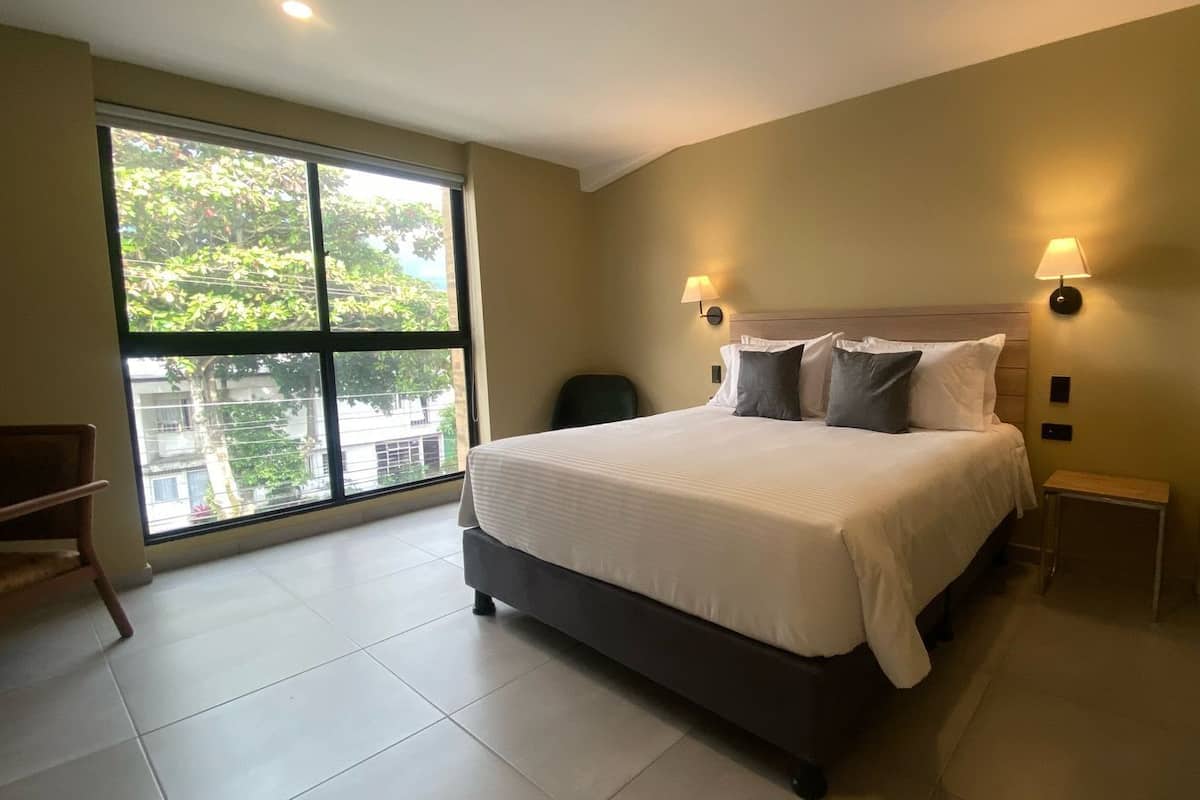 A modern bedroom with a large bed, two bedside lamps, a view through a large window, and a small seating area.