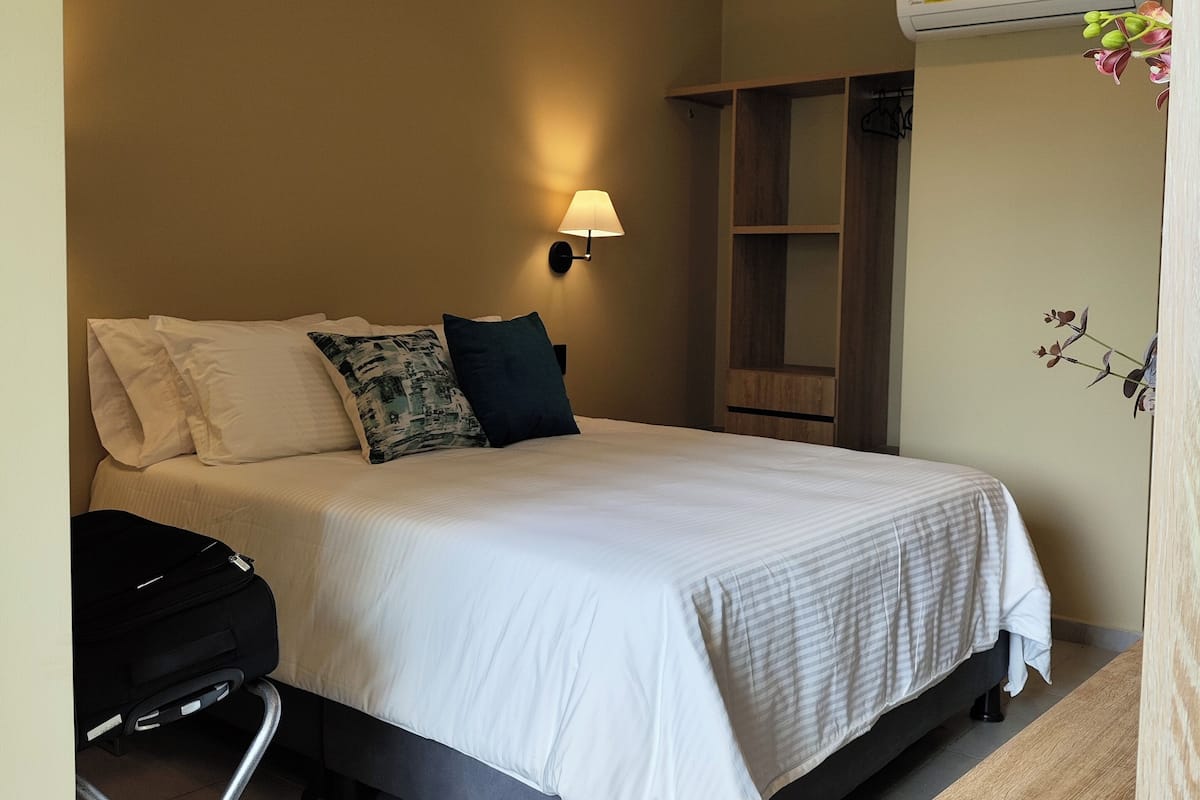 A hotel room with a bed, a bedside lamp, a suitcase, a wooden wardrobe, and an air conditioning unit.