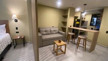 A compact hotel room with a bed, a sofa, a small table, a desk with stools, and a kitchenette.