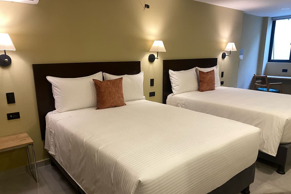 Two double beds with white linens and brown pillows, a small bedside table, and wall-mounted lamps.