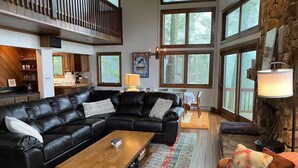 A spacious living room with a large black leather sectional sofa, a wooden coffee table, and a colorful rug. There are multiple windows providing natural light and a stone wall accentuating the space. - Slopes @ Tyro (Wintergreen Resort)