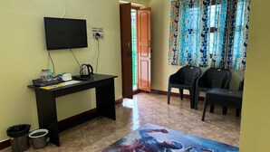 Une pièce avec un téléviseur fixé au mur, un bureau avec une bouilloire et des bouteilles d’eau, deux poubelles et un tapis coloré sur le thème des super-héros.