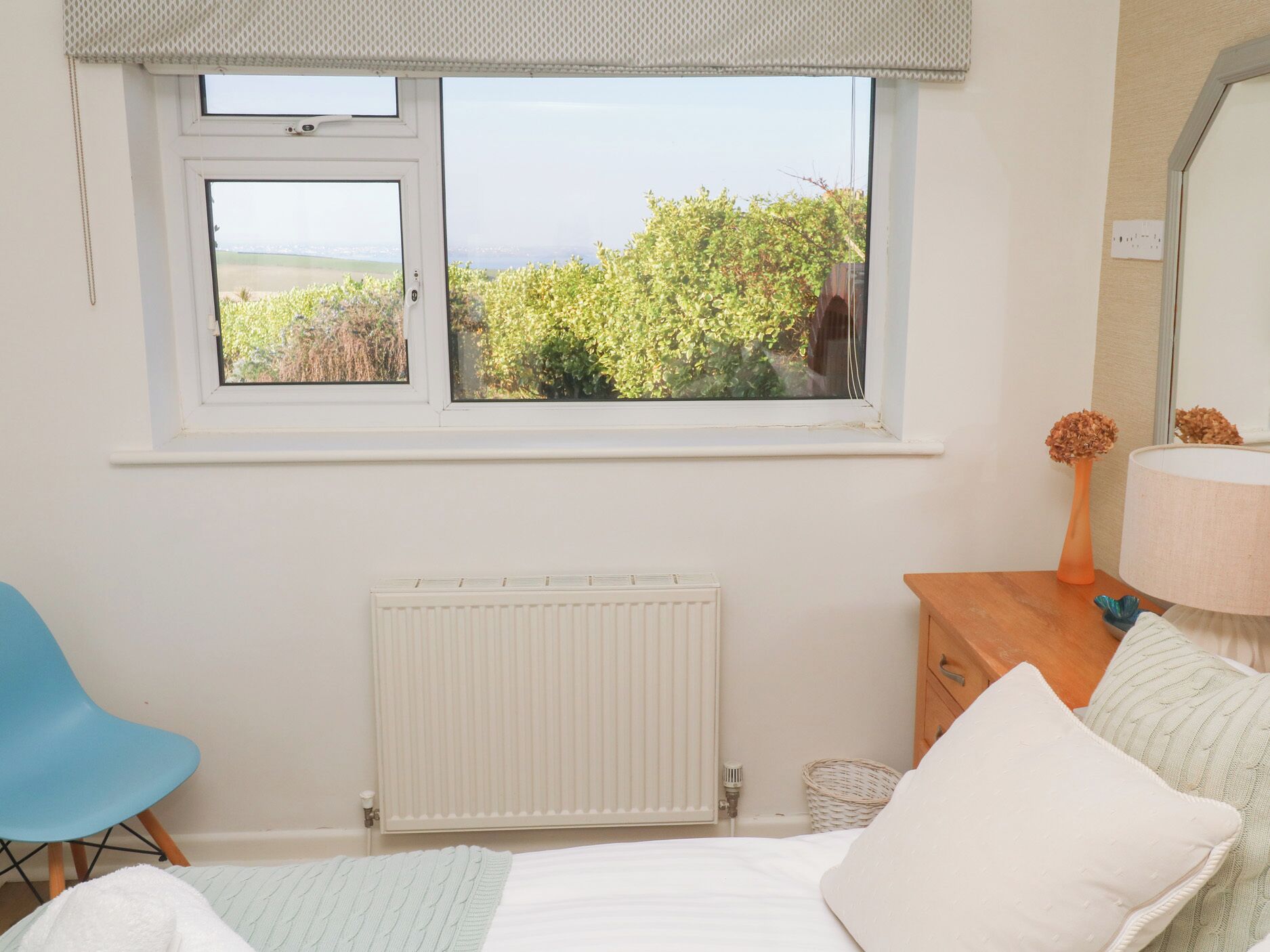 Una camera da letto con una grande finestra, un termosifone, una sedia blu, un comò in legno e una lampada.
