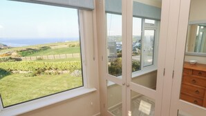 A room with a large window offering a view of a green landscape and a distant body of water. - Coastman's Nest (Newquay)