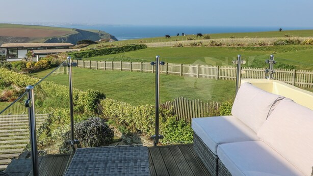 A deck with a white sofa and a view of a grassy field with cows, a wooden fence, and a building in the background. - Coastman's Nest (Newquay)